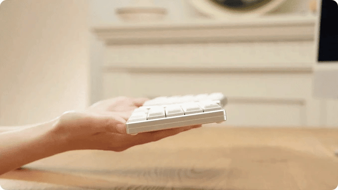 A lifestyle photograph showing a person's hand holding one silver half of the ElimKeys Elytra split mechanical keyboard from a side angle. The shot emphasizes the keyboard's extremely thin, low-profile design, featuring a sleek aluminum chassis and white low-profile keycaps against a warm, blurred interior background.